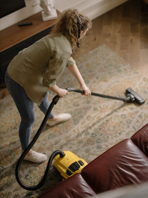Close-up of a modern vacuum cleaner with a clear dust container filled with debris, connected to a flexible, patterned hose attached to a black cleaning head on a light pink carpeted floor. The vacuum