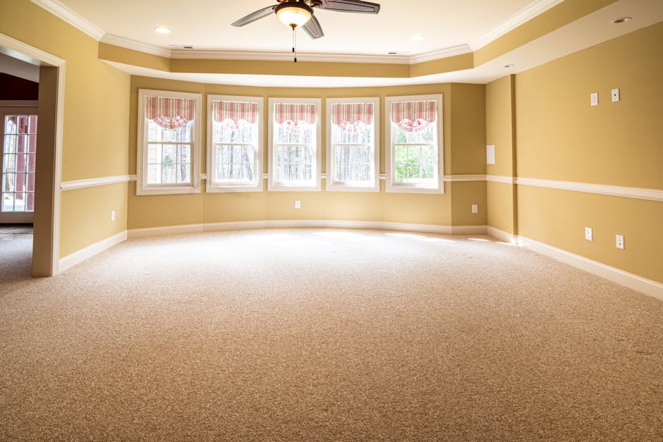 A spacious, well-lit living room with a neutral beige carpet that appears clean and plush, along with smooth, pale yellow walls accented by a white chair rail. The room features a white ceiling fan wi