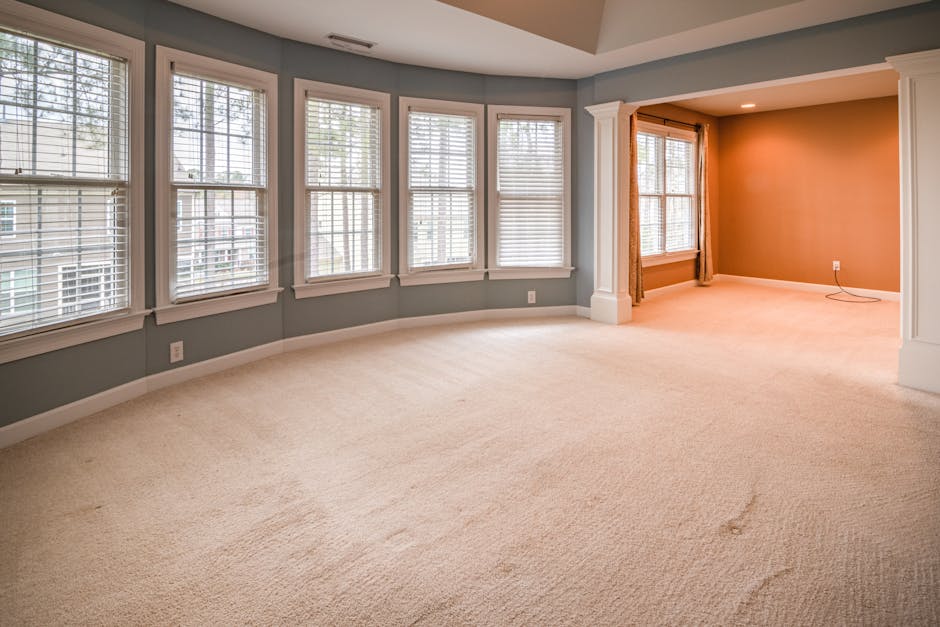 A spacious, well-lit living room with a neutral beige carpet that appears clean and plush, along with smooth, pale yellow walls accented by a white chair rail. The room features a white ceiling fan wi
